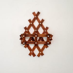 May include: A brown wooden wall-mounted rack featuring a diamond-shaped lattice structure with a horizontal shelf section. The wooden slats have a ribbed texture and are connected by small, round, white decorative accents at each intersection.