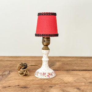 May include: A small, vintage table lamp with a red lampshade trimmed with black. The lamp has a white ceramic base decorated with floral patterns and a brass bulb socket. The lamp is on a wooden surface.