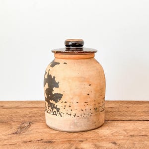 May include: A vintage ceramic jar with a lid. The jar has a rounded body and is light tan with dark brown abstract patterns. The lid is dark brown. The jar sits on a wooden surface.