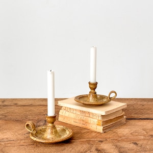 Puede incluir: Dos candelabros de cerámica con velas blancas, uno sobre una pila de libros. Los candelabros están moteados con un asa lateral. El fondo es una pared blanca lisa y una mesa de madera.