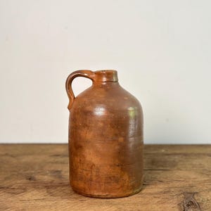 May include: A brown stoneware jug with a handle. The jug has a rounded body and a narrow neck. The surface has a mottled appearance. The jug is sitting on a wooden surface.