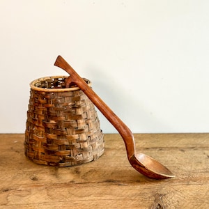 May include: A brown woven basket with a tapered cylindrical shape and a long-handled wooden spoon resting against it. The spoon features a smooth, polished finish and a curved handle, while the basket displays a complex, interlaced pattern.