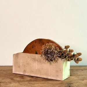 Peut inclure: Une boîte en bois rectangulaire et rustique, avec un dos en bois incurvé. La boîte est peinte en rose et vert délavés. Des fleurs séchées et des gousses sont disposées à l'intérieur, créant une décoration naturelle.