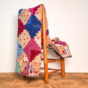 May include: A patchwork quilt featuring a log cabin pattern in shades of magenta, blue, and beige, accented with small red yarn ties throughout. The quilt is draped over a light brown wooden chair with four legs and horizontal support rungs.