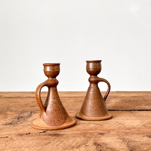 Puede incluir: Dos candeleros de cerámica marrón con asa. Los candeleros tienen un diseño moteado y una base ancha. El fondo es una pared blanca y una mesa de madera.