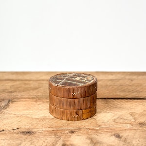 May include: A small, round, wooden box with a lid. The box is made of stacked, textured wood and has a dark brown top with a grid-like pattern. The box sits on a wooden surface.