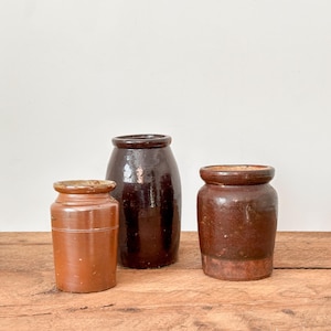 May include: Three antique stoneware jars in varying sizes and shades of brown. The jars are cylindrical with rounded shoulders and rims. The jars are displayed on a wooden surface against a white background. The jars are likely for storage.