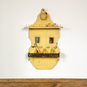 Puede incluir: Un organizador de costura de madera amarilla vintage con un acabado envejecido. Cuenta con un estante con carretes y agujas, un compartimento inferior y una barra de metal. El organizador está montado en una pared blanca.