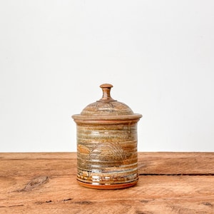 May include: A handmade ceramic jar with a lid. The jar has a cylindrical form with a textured surface and a lid with a small knob. The colour scheme includes shades of brown, tan, and orange, creating a rustic aesthetic. The jar is likely used for storage.
