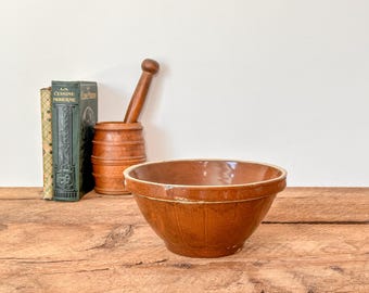 Puede incluir: Un cuenco de cerámica marrón con un borde marrón claro se encuentra sobre una superficie de madera. Junto a él hay dos libros antiguos y un mortero y una maja de madera. El cuenco tiene un diseño clásico y rústico.