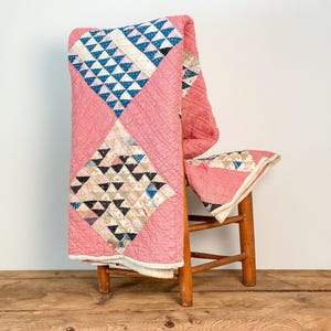 Puede incluir: Un edredón vintage sobre una silla de madera. El edredón presenta un fondo rosa con patrones de diamantes de triángulos azules, negros y beige. El edredón está hecho de tela suave y tiene un diseño acolchado.