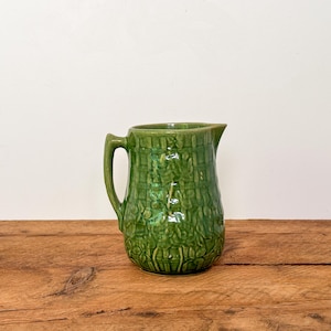 Peut inclure: Une cruche en céramique verte avec une surface texturée et une anse. La cruche a un bec verseur et est posée sur un fond blanc sur une surface en bois. La cruche est d'une nuance de vert vif.
