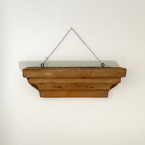 May include: A wooden wall shelf with a layered design, suspended by wire. The shelf is a warm brown colour and is mounted on a white wall. The shelf is likely for displaying decorative items.