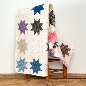 May include: A cream-coloured quilt with a star pattern in various colours, including blue, purple, and grey, draped over a wooden chair. The quilt's design features eight-pointed stars, adding a touch of vintage charm to the scene.