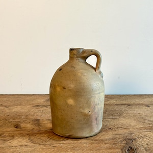 May include: A vintage stoneware jug with a handle. The jug is a light brown colour with a rough, textured surface. It has a slightly rounded body and a narrow neck.