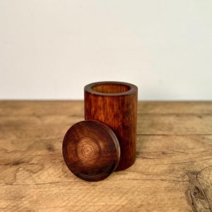 Könnte beinhalten: Ein zylindrischer Holzbehälter mit passendem kreisförmigen Deckel. Der Behälter und der Deckel haben eine satte, warme braune Farbe mit sichtbarer Holzmaserung. Der Deckel liegt neben dem Behälter und gibt den Blick ins Innere frei.