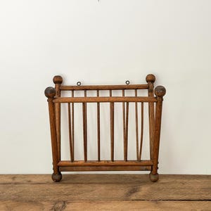Puede incluir: Un revistero de madera vintage con un acabado marrón cálido. El revistero presenta listones verticales y remates redondeados en las esquinas. Está diseñado para montaje en pared y se apoya sobre una superficie de madera.