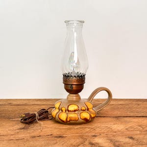 May include: A ceramic oil-style lamp featuring a rounded base with a textured yellow and brown glaze, a curved handle, and a copper-colored metal collar. The lamp is topped with a clear glass chimney and includes a brown electrical cord and plug.