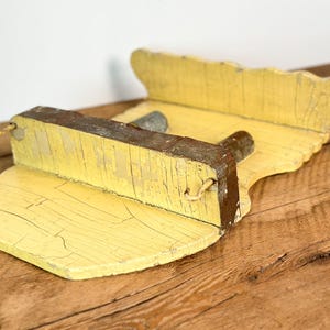 K&ouml;nnte beinhalten: Ein verwittertes, gelbes Holzregal. Das Regal hat eine gewellte R&uuml;ckseite und einen rechteckigen Vorderteil. Das Holz weist Gebrauchsspuren und Risse auf, die auf das Alter hindeuten. Ein kleines Metallst&uuml;ck ist am Regal befestigt.