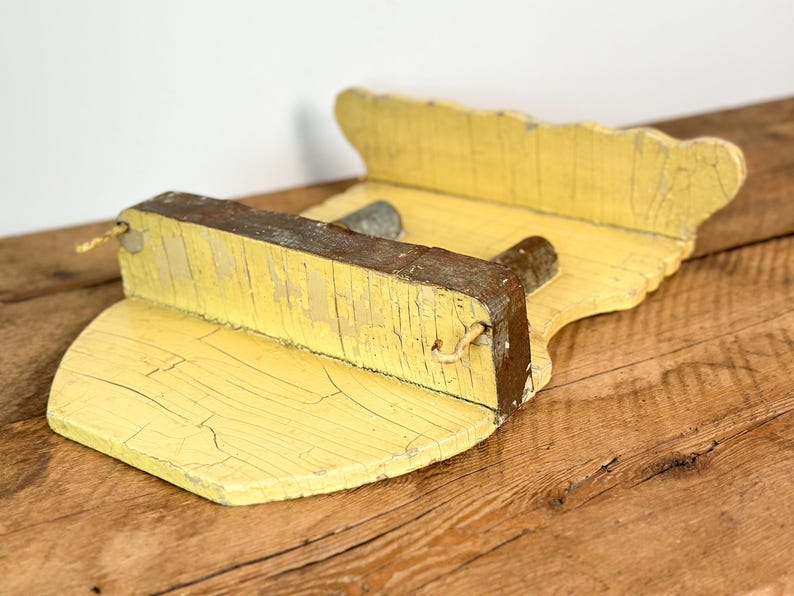 K&ouml;nnte beinhalten: Ein verwittertes, gelbes Holzregal. Das Regal hat eine gewellte R&uuml;ckseite und einen rechteckigen Vorderteil. Das Holz weist Gebrauchsspuren und Risse auf, die auf das Alter hindeuten. Ein kleines Metallst&uuml;ck ist am Regal befestigt.