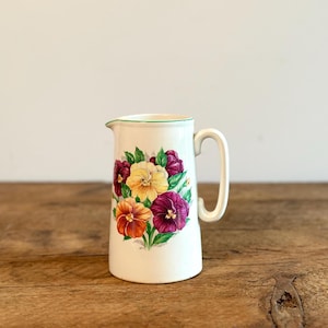 May include: White ceramic pitcher with a green rim and a floral design featuring purple, yellow, and orange pansies.