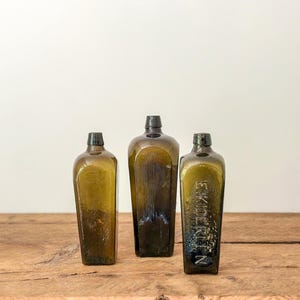 Puede incluir: Tres botellas antiguas de vidrio verde oliva con tapones oscuros. Las botellas son rectangulares con hombros redondeados y están sobre una superficie de madera. Una botella tiene el texto en relieve "EKIDEKTIN".