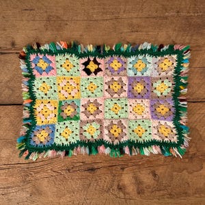 Peut inclure: Une couverture au crochet faite à la main, colorée, avec une frange verte. La couverture présente un motif patchwork avec des carrés aux teintes pastel de rose, jaune, bleu et vert. La couverture est présentée sur une surface en bois rustique.