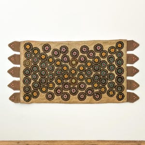 May include: A rectangular textile with a tan background and numerous embroidered circles. Each circle features a floral design with yellow, pink, and blue accents. The edges have brown, decorative tabs.