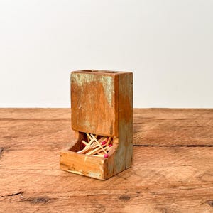 May include: A rustic wooden match holder with a distressed finish. The holder is rectangular with a square opening at the top and a lower compartment filled with matches. The wood has a warm, natural tone with hints of green paint.