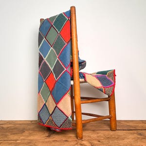 May include: A wooden chair draped with a blanket featuring a diamond-shaped patchwork pattern in various colours including blue, red, green, and tan. The fabric segments are joined by visible stitching, creating a geometric design across the textile.