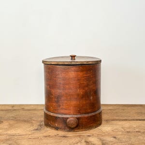May include: A cylindrical wooden container with a lid and a small knob on top. The container is a warm brown color and sits on a wooden surface. A wooden knob is on the side of the container.