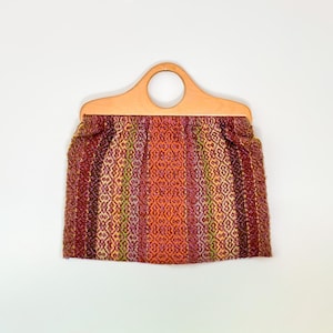 Puede incluir: Bolso de mano vintage con asa de madera y cuerpo de tela tejida. La tela presenta rayas verticales en tonos rojos, naranjas y verdes. El asa tiene una abertura circular.