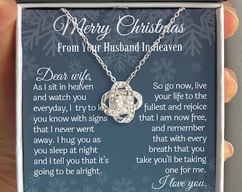 May include: A silver-toned necklace with a central knot pendant featuring a clear crystal, presented in a black box with a blue card. The card displays the text Merry Christmas From Your Husband In Heaven and a message to a wife about love and comfort.