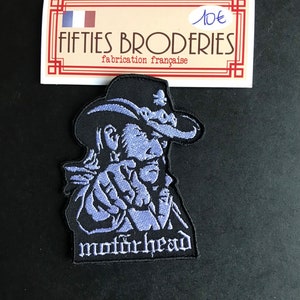 Pode incluir: Um remendo bordado preto e roxo com uma imagem de desenho animado de um homem usando um chapéu de cowboy e o texto "motorhead".