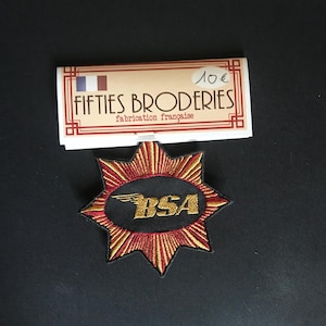 Peut inclure: Un écusson brodé en forme d'étoile avec un centre ovale noir portant le texte doré BSA et un logo ailé. Les pointes de l'étoile sont cousues avec du fil rouge et doré. L'écusson est fixé sur une carte rectangulaire marquée FIFTIES BRODERIES.