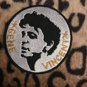 Pode incluir: Um remendo redondo bordado em preto e branco com contorno prateado. O remendo apresenta uma silhueta da cabeça de uma pessoa com o texto "GENE" e "VINCENT" em letras douradas.