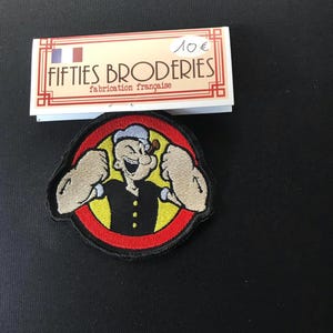 May include: A circular embroidered patch featuring a cartoon character with large muscular arms, a black shirt, and a white sailor hat against a yellow and red background. The patch is attached to a white card labeled FIFTIES BRODERIES.