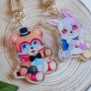 Puede incluir: Dos llaveros de peluche, uno es un oso rosa y azul que lleva un sombrero de copa negro y sostiene un micrófono, el otro es un conejo blanco con la nariz rosa y un lazo azul que sostiene un cuchillo rosa.