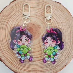 May include: Two clear acrylic keychains featuring a cartoon character with purple hair and a pink bow, hugging a green creature. The keychains have a silver clasp and are on a wooden surface. The text "Boo Erne" is visible.