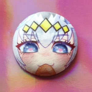 May include: A circular plush toy with an anime-style face, large blue eyes, pink details, and a yellow and white crown. The plushie has a soft texture and is set against a pink background.