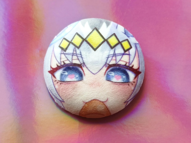 May include: A circular plush toy with an anime-style face, large blue eyes, pink details, and a yellow and white crown. The plushie has a soft texture and is set against a pink background.