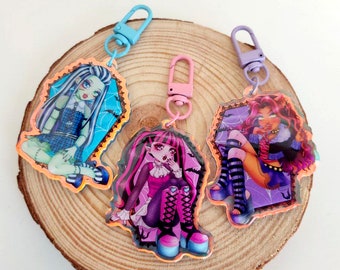 May include: Three colourful keychains featuring cartoon characters from the Monster High franchise. The keychains are made of clear acrylic with a glossy finish. The characters are Frankie Stein, Draculaura, and Lagoona Blue. Each keychain has a metal ring and a lobster clasp.