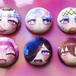 May include: Six circular plush badges featuring anime-style character faces. Each badge displays a different character with unique expressions and colour schemes, including blue, pink, and brown hair. The badges are set against a pink background.