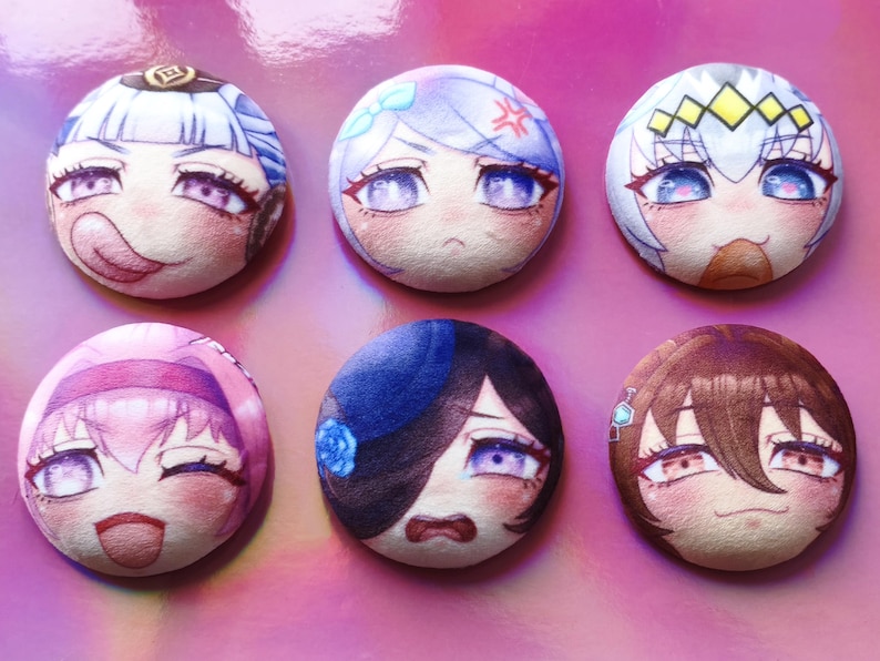 May include: Six circular plush badges featuring anime-style character faces. Each badge displays a different character with unique expressions and colour schemes, including blue, pink, and brown hair. The badges are set against a pink background.