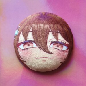 May include: A circular plush toy with an anime-style face, brown hair, red eyes, and rosy cheeks. A small, light blue gem is visible on the side. The plushie is set against a pink background.