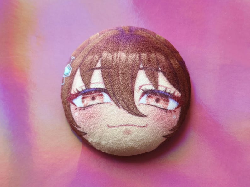 May include: A circular plush toy with an anime-style face, brown hair, red eyes, and rosy cheeks. A small, light blue gem is visible on the side. The plushie is set against a pink background.