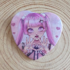 May include: A pink heart-shaped button with a cartoon girl with pink hair and a pink and white outfit. The text on the button reads "I hate myself and I want to die".