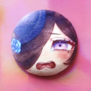 May include: A circular plush toy with an anime-style face, featuring purple eyes, dark hair, and a blue hat with a flower. The face has a distressed expression with a red mouth and blush. The background is a pink gradient.