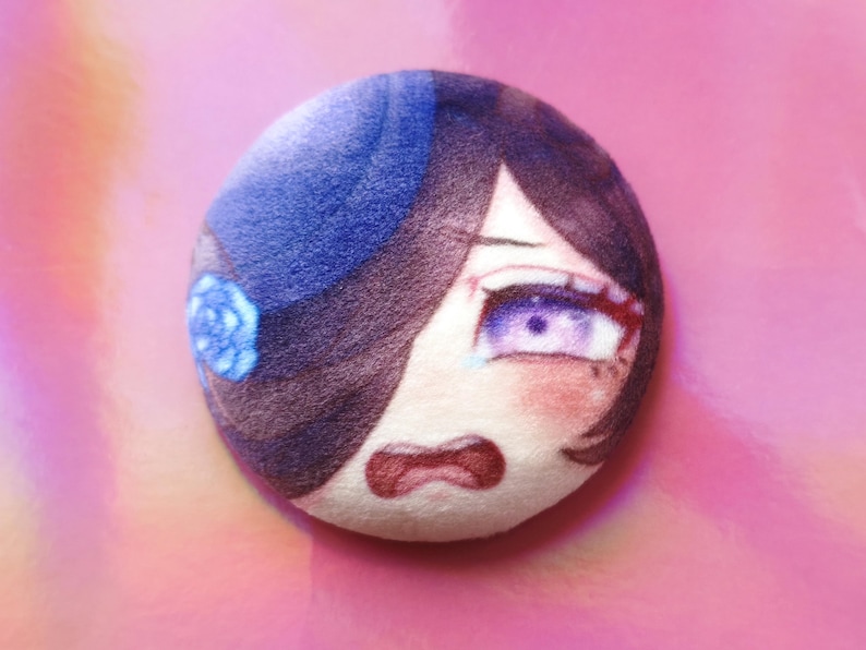 May include: A circular plush toy with an anime-style face, featuring purple eyes, dark hair, and a blue hat with a flower. The face has a distressed expression with a red mouth and blush. The background is a pink gradient.