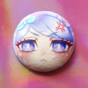 May include: A circular badge displaying an anime-style character's face with purple eyes, light blue hair, and a red angry symbol. The badge has a soft, textured appearance and a light pink background.
